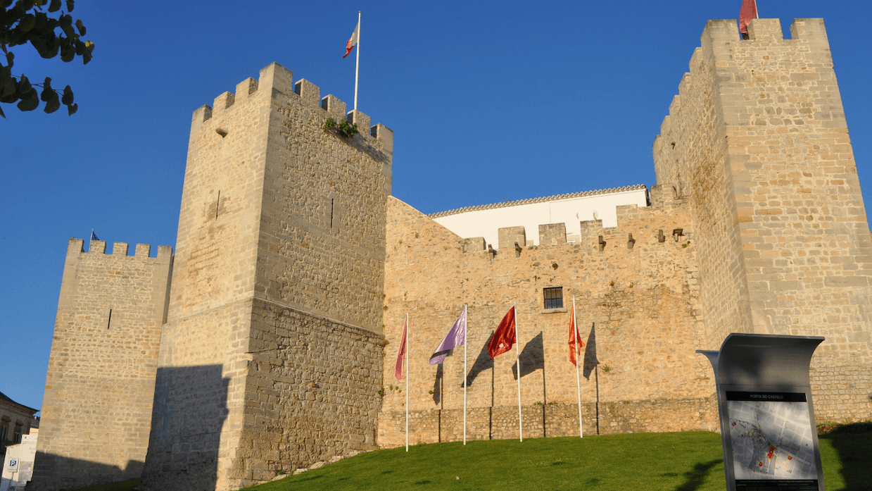 A stone castle with towers and battlements, surrounded by flags on poles, under a clear blue sky.