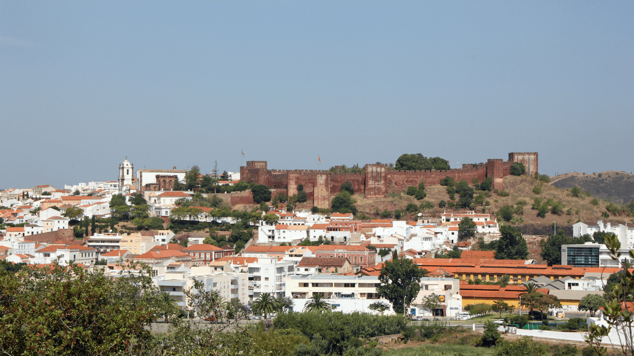 A panoramic view of Silves, Portugal, featuring white buildings, a historic red castle, and a church under a clear blue sky.