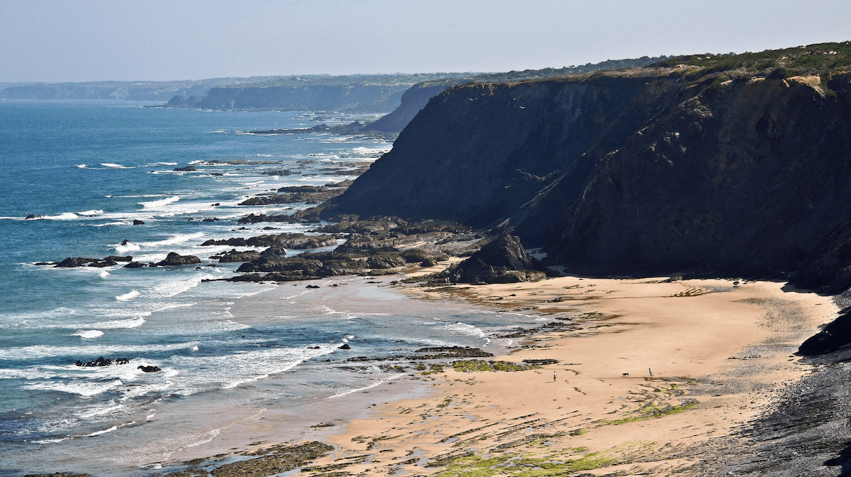 Felsige Küstenlinie mit steilen Klippen, sandiger Strand und Wellen, die unter einem klaren blauen Himmel gegen das Ufer schlagen.