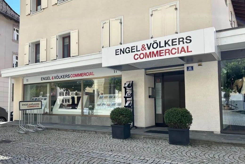 Storefront of Engel & Völkers Commercial with large windows displaying images, next to a tree-lined cobblestone street.