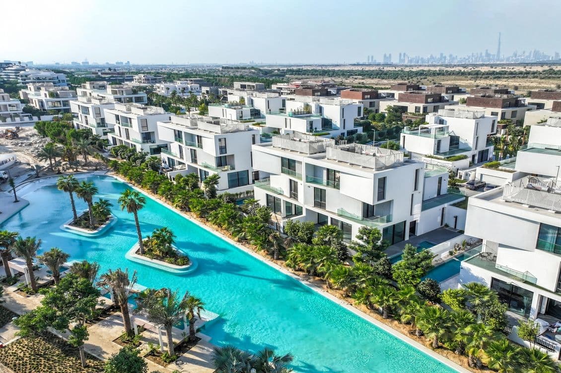 Luftaufnahme moderner weißer Villen mit Flachdächern, umgeben von üppigem Grün und einem langen, gewundenen türkisfarbenen Pool, vor der Kulisse einer fernen Stadt-Skyline.