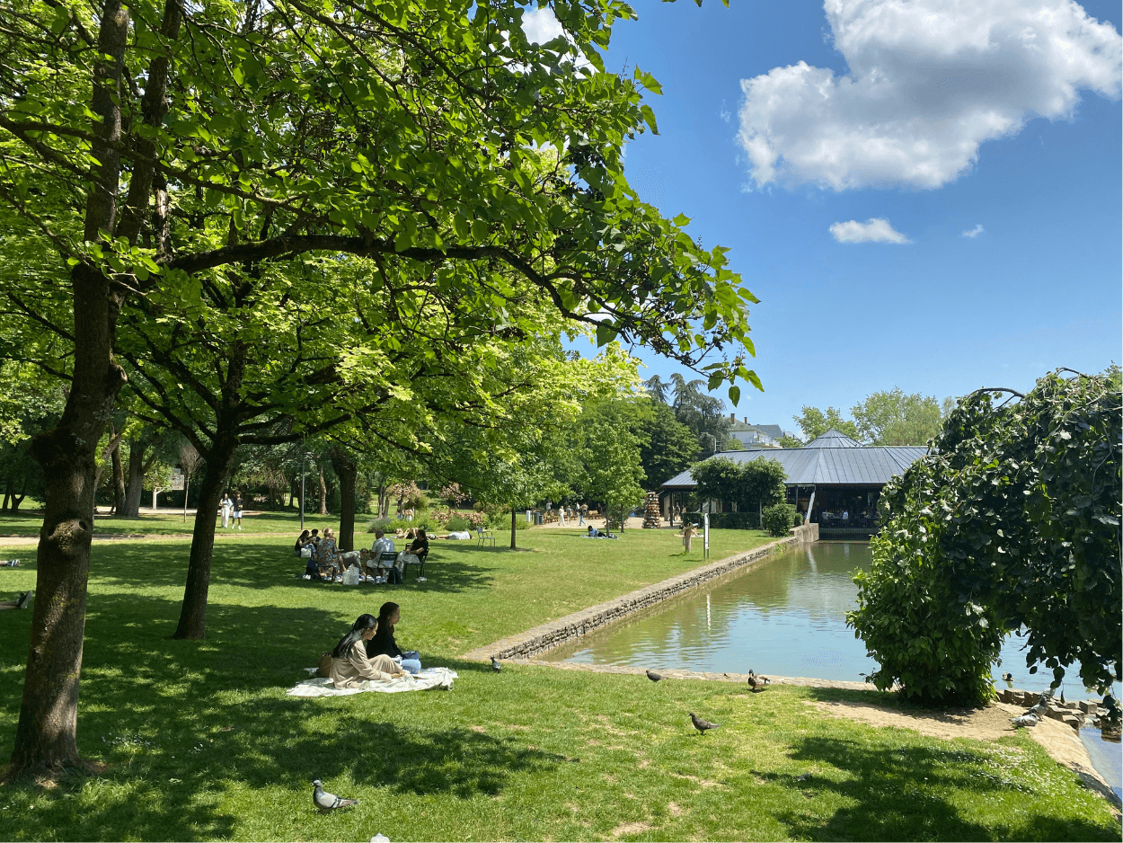 Des gens se détendent dans un parc par une journée ensoleillée. Un étang est visible avec un bâtiment en arrière-plan.
