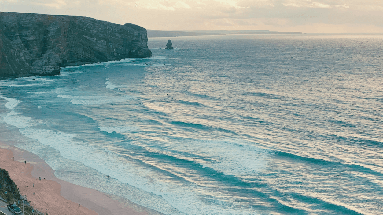Coastal landscape with cliffs, sandy beach, and gentle waves under a cloudy sky. People walk along the shore.