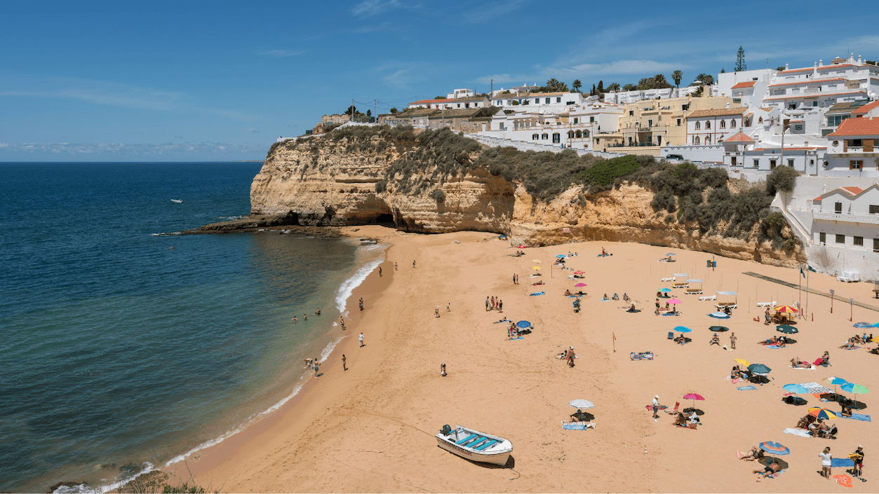 Sunny beach with people, colorful umbrellas, and a boat on the sand. Cliffs and white buildings overlook the ocean under a clear blue sky.