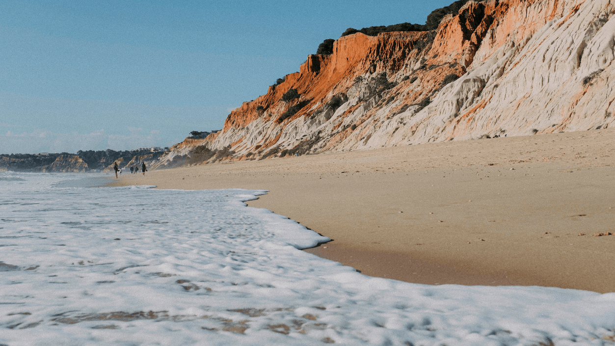 Waves gently wash onto a sandy beach with tall, colorful cliffs in the background under a clear blue sky.
