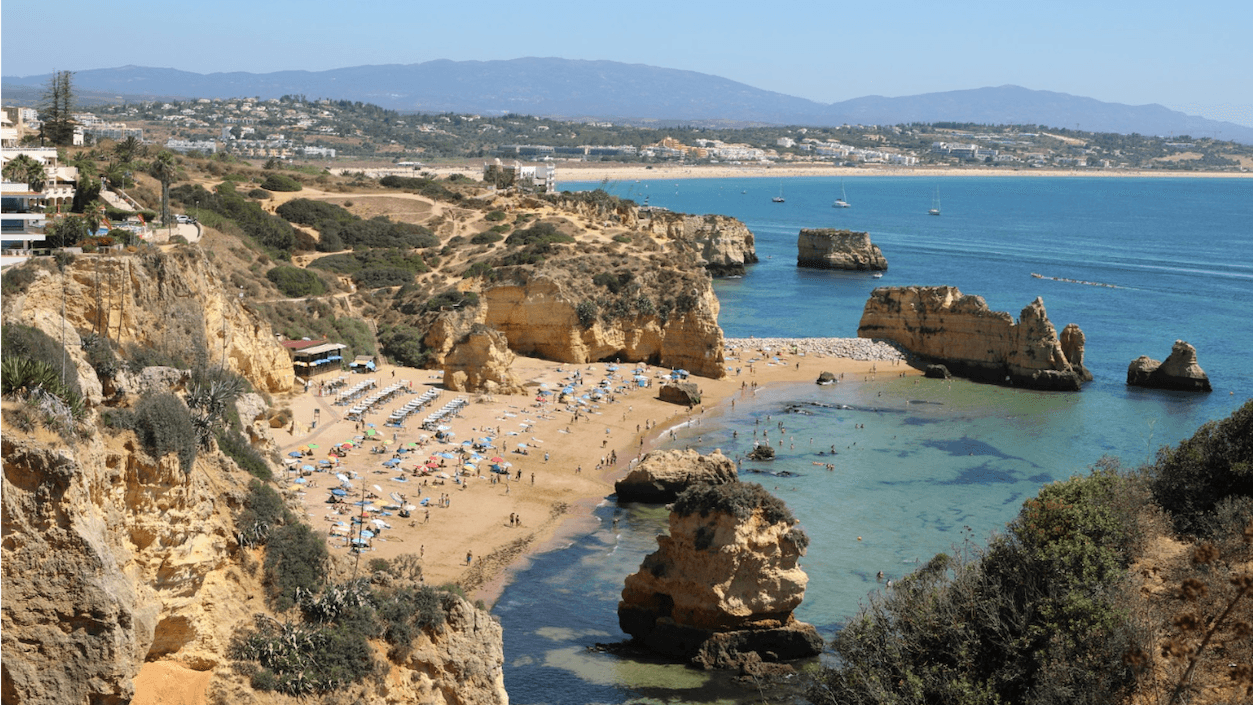 Scenic view of a sandy beach with sunbathers, colorful umbrellas, rocky cliffs, and clear blue sea under a sunny sky.