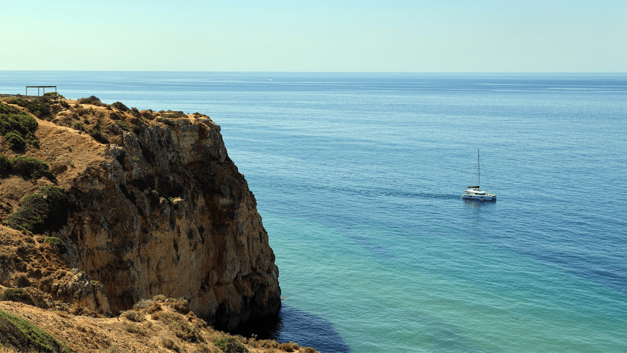 A sailboat on calm blue waters near a rugged cliff, with a clear sky above and sparse vegetation on the cliffside.
