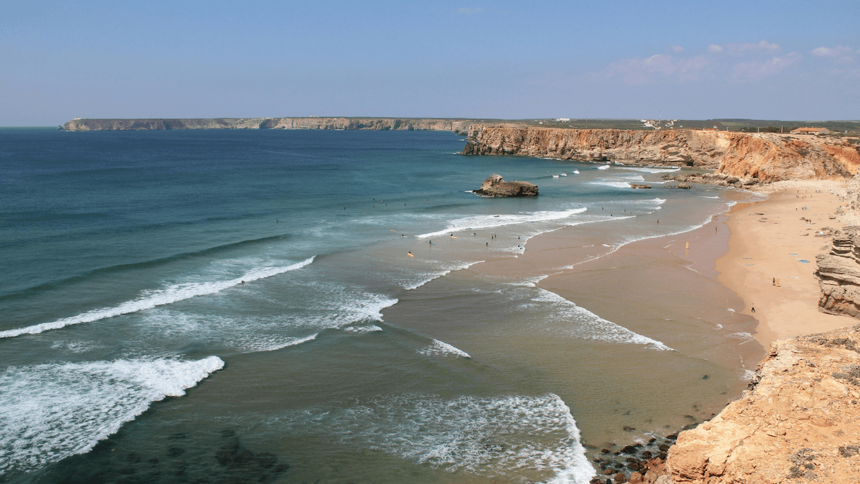 A scenic view of a coastal beach with turquoise waves, sandy shores, and rocky cliffs under a clear blue sky.