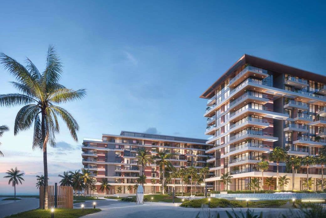 Modern beachfront residential buildings at dusk, featuring wide balconies, warm exterior lighting, palm trees, landscaped walkways, and an ocean backdrop under a clear evening sky.
