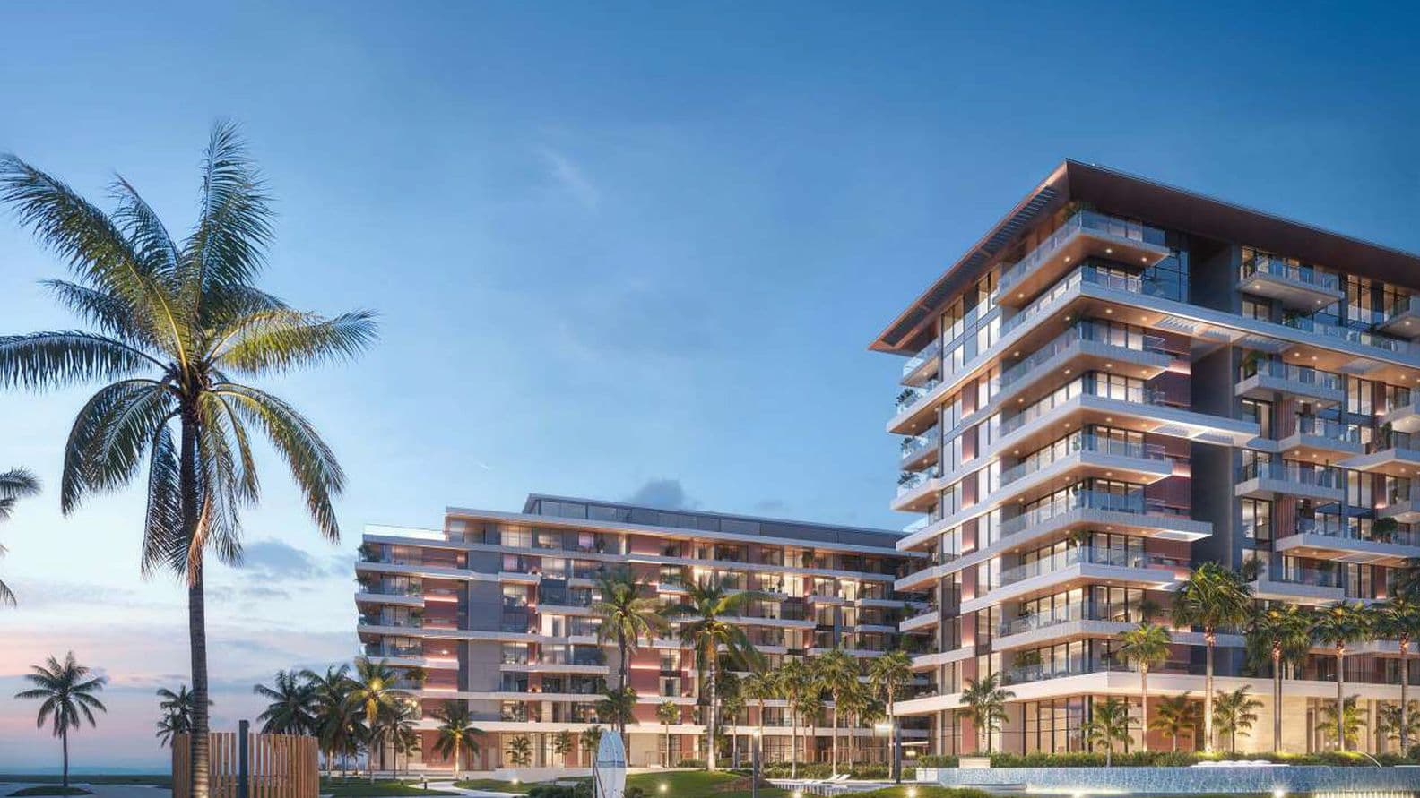 Modern beachfront residential buildings at dusk, featuring wide balconies, warm exterior lighting, palm trees, landscaped walkways, and an ocean backdrop under a clear evening sky.