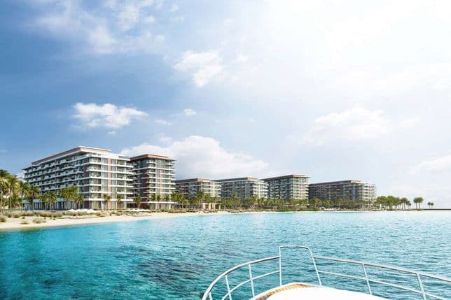 View from a boat approaching a row of modern beachfront residential buildings, with clear turquoise water, sandy shore, palm trees, and a bright sky overhead.