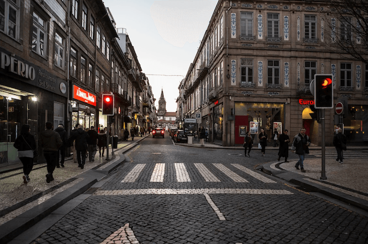 A cobblestone street with a zebra crossing, flanked by historic buildings, people walking, and a red traffic light at dusk.