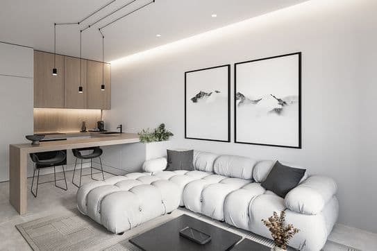Modern open-plan living area with a tufted white sofa, light wood kitchen island, two black-framed mountain prints, and pendant lighting.