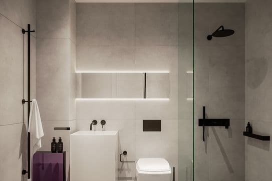 Minimalist gray bathroom: wall‑mounted toilet, white pedestal sink, glass shower panel, black fixtures and a purple side table.