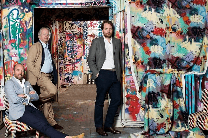 Trois hommes posent dans une pièce recouverte de graffitis. L'un est assis dans un fauteuil à motifs, un autre est appuyé contre une porte, et le troisième se tient debout, la main dans la poche.