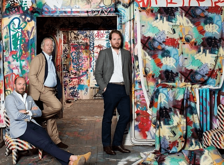 Three men pose in a graffiti-covered room. One sits in a patterned chair, another leans against a doorway, and the third stands with his hand in his pocket.
