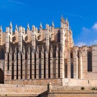 Gotische Kathedrale mit filigranen Türmen unter blauem Himmel, ein Brunnen links im Bild, umgeben von historischen Gebäuden und Bäumen.