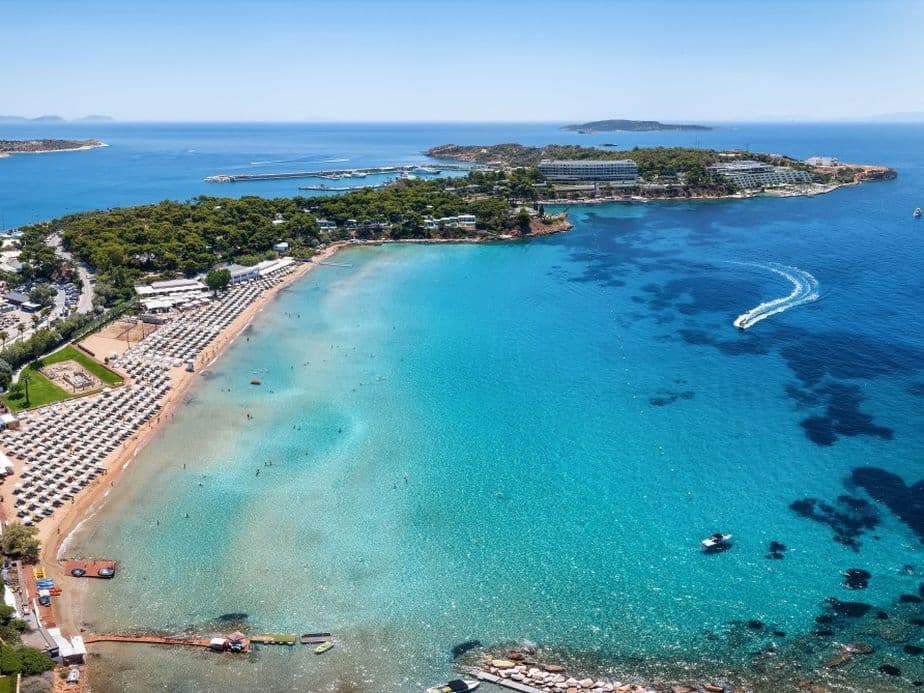 Aerial view of Astir Beach, Greece, with turquoise water, sandy beach, rows of umbrellas, and boats on the blue sea.