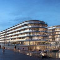 Futuristic waterfront buildings with curved glass facades at sunset, surrounded by boats and palm trees under a clear blue sky.