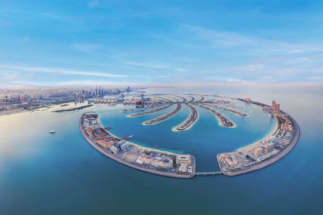Aerial view of aPalm Jumeirah with surrounding water, buildings, and a distant city skyline under a clear blue sky.
