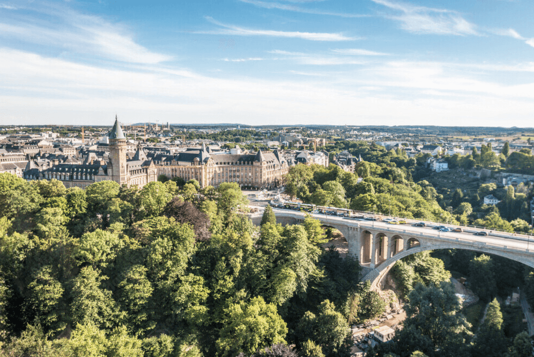 Luxembourg City view with a stone bridge filled with cars, surrounded by green trees and buildings under a blue sky.
