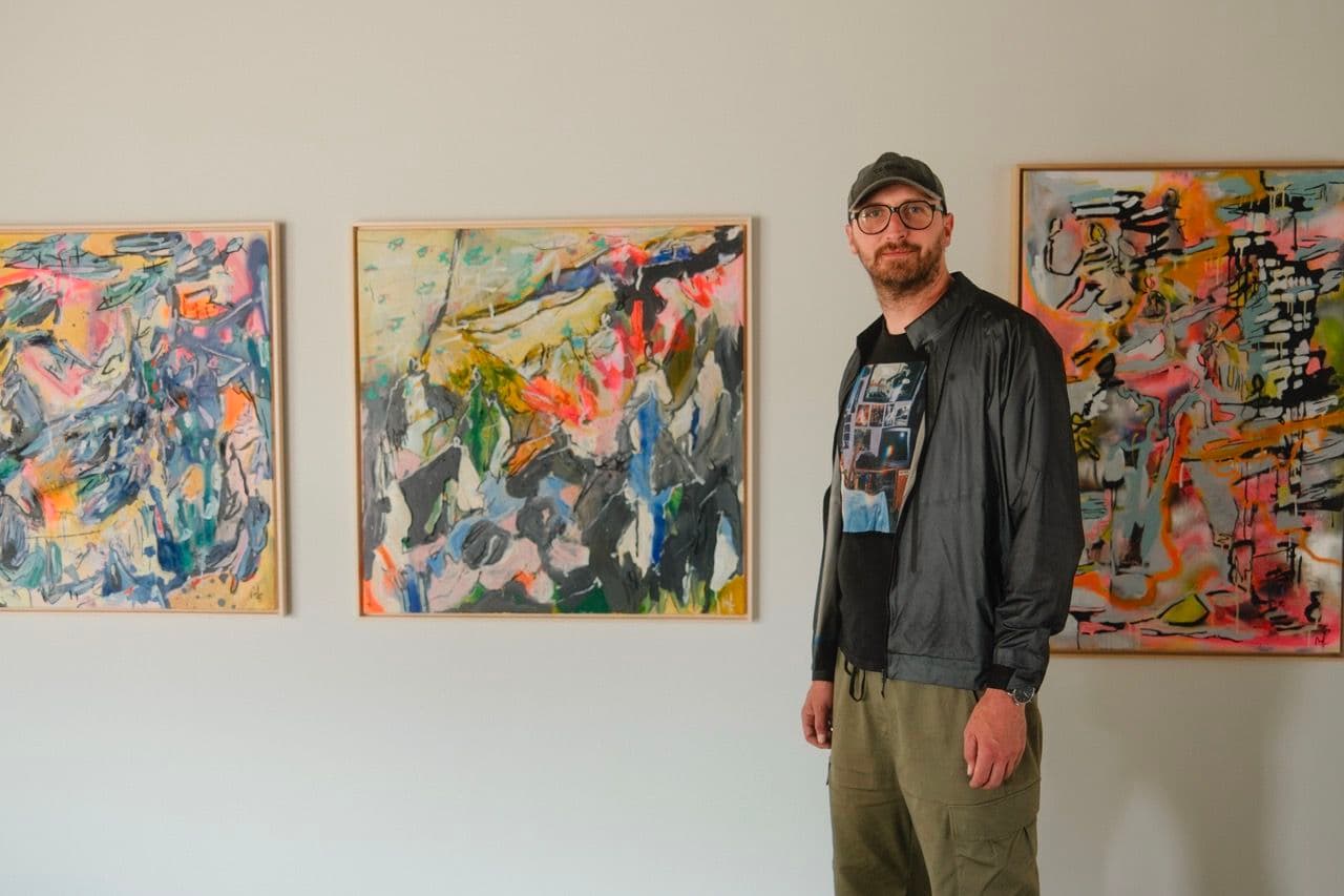 A person stands in a gallery in front of three abstract, colorful paintings hanging in wooden frames on a white wall.