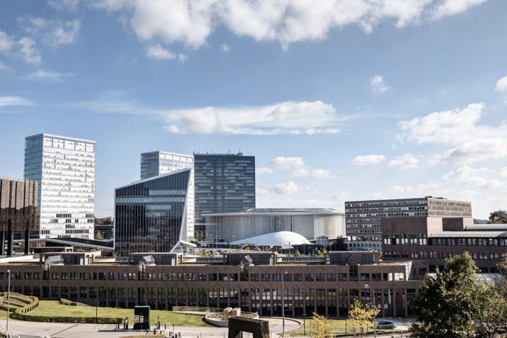 Stadtbild mit modernen Gebäuden unter blauem Himmel mit vereinzelten Wolken. Die Gebäude haben unterschiedliche Höhen und architektonische Stile.