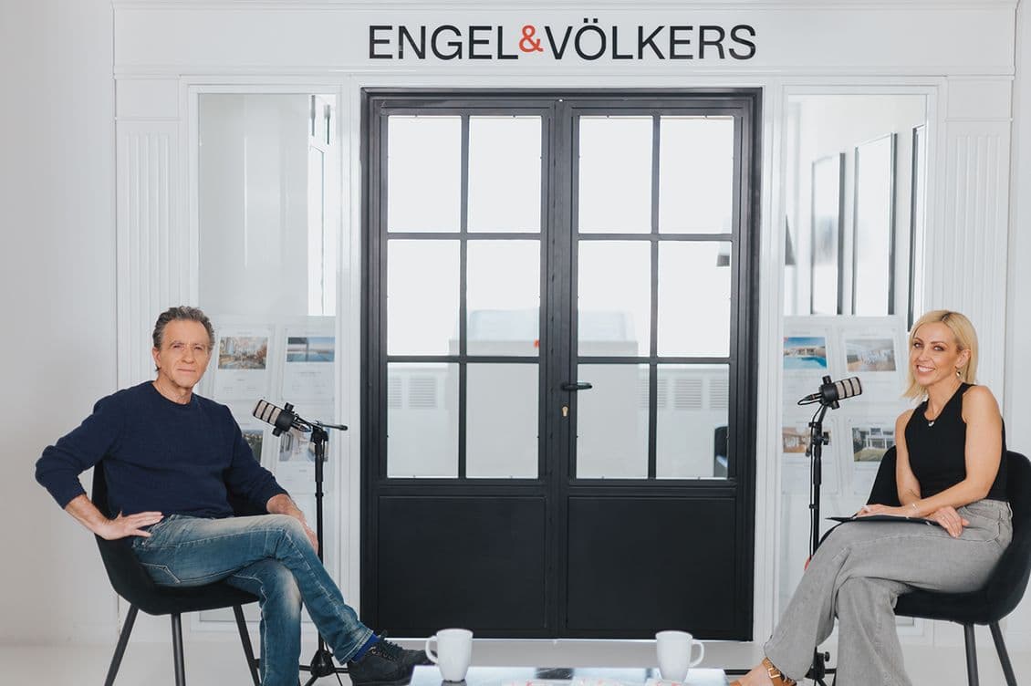 Κώστας Μπίγαλης και Λίλη Πυράκη κάθονται σε καρέκλες με μικρόφωνα ανάμεσά τους, έτοιμοι για ένα podcast ή μια συνέντευξη σε ένα μοντέρνο, φωτεινό γραφείο. Η ένδειξη ENGEL & VÖLKERS αναγράφεται πάνω από γυάλινες πόρτες πίσω τους, κάνοντας podcast.