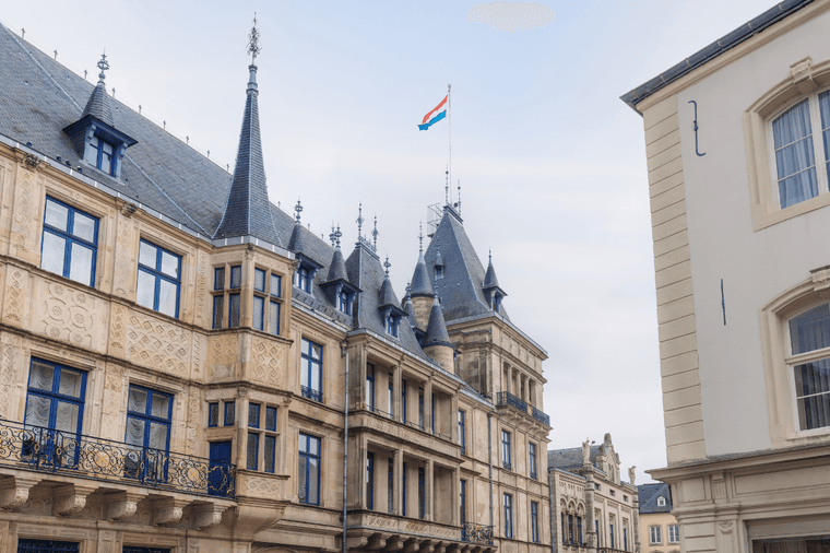 Bâtiment beige orné avec des fenêtres bleues et un drapeau luxembourgeois au sommet sur fond de ciel nuageux.
