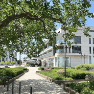 Une vue ensoleillée d'un complexe d'appartements moderne avec une allée en pierre, des arbres verts et des parterres de fleurs.