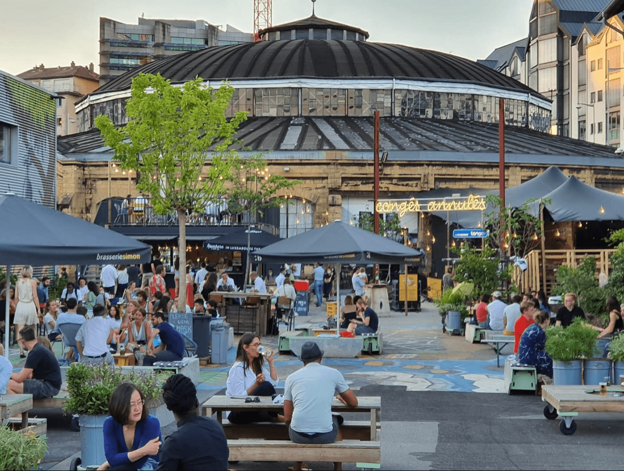 Outdoor market scene with people seated at tables under tents. A large, round building with a domed roof is in the background. The sky is dusky.