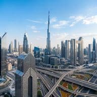 Skyline of a modern city with skyscrapers, including a tall iconic tower, surrounded by roads and highways under a clear blue sky.