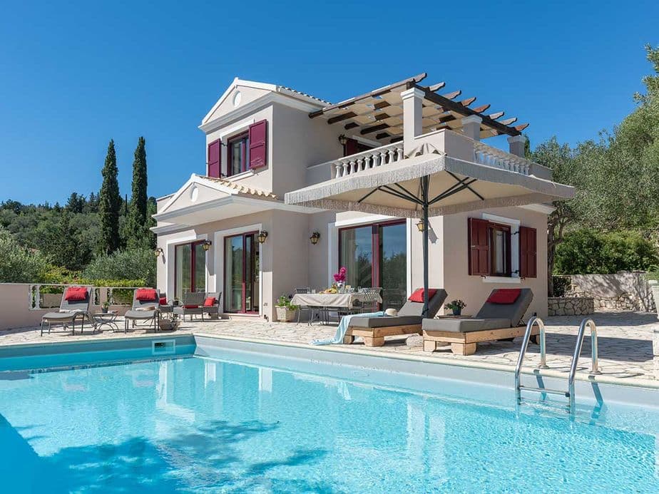 A modern two-story villa with red shutters, a pergola, and lounge chairs by a clear blue swimming pool, surrounded by lush greenery under a bright blue sky.