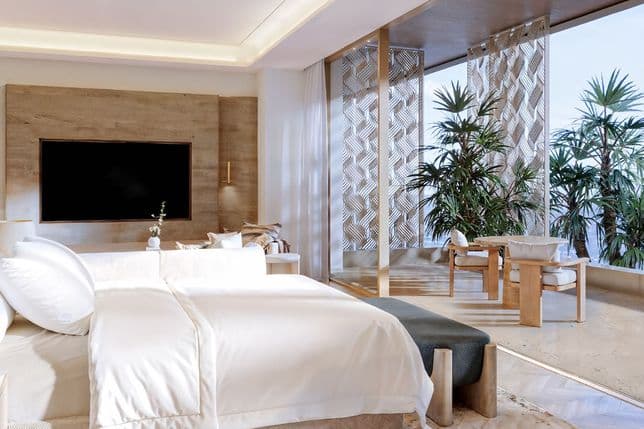 Luxurious bedroom with a large bed, fluffy white bedding, wooden accents, floor-to-ceiling windows, and indoor plants, under soft ceiling lights.