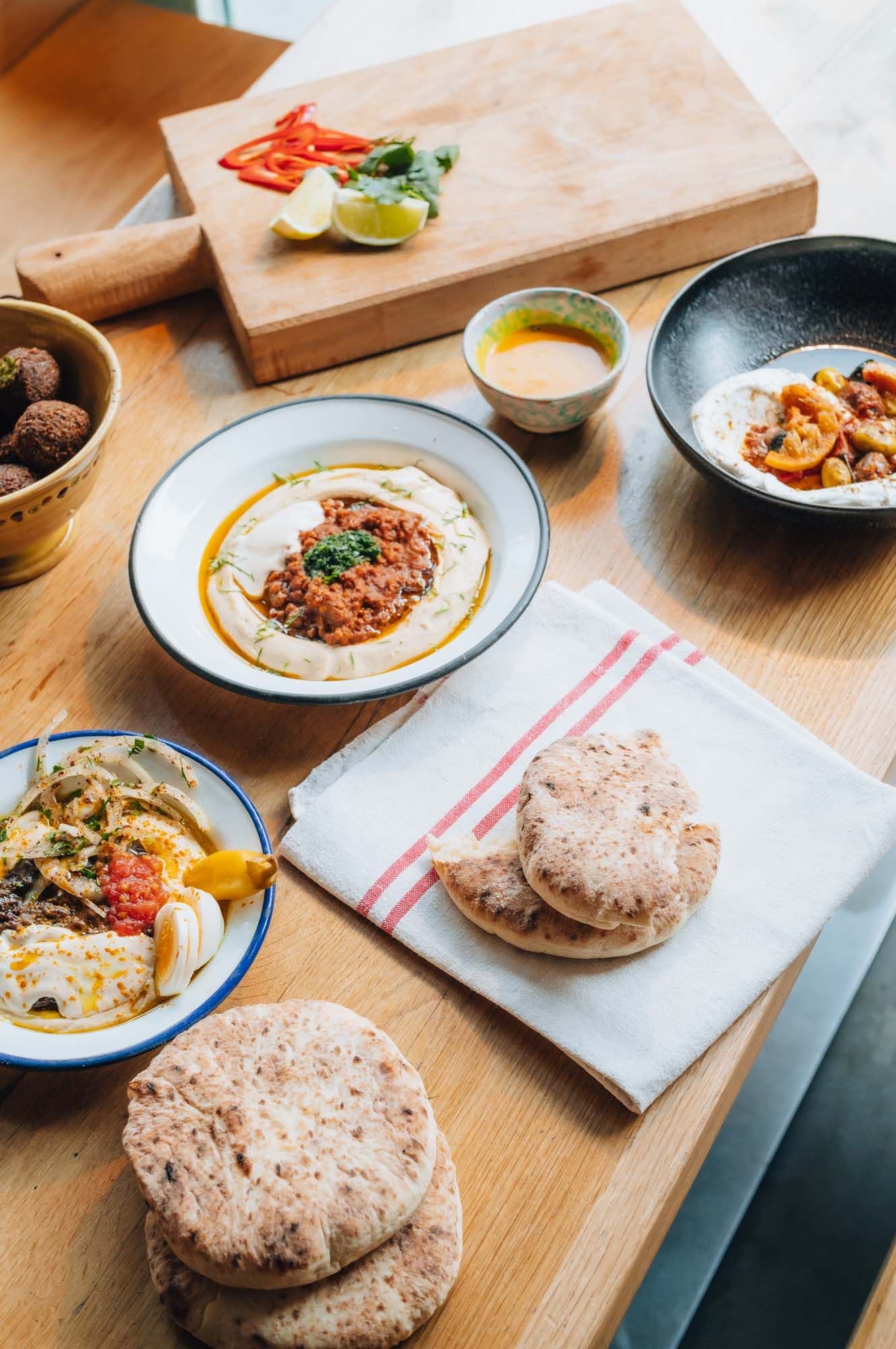 A wooden table with plates of hummus, falafel, pita bread, and a cutting board with lime and sliced red peppers.
