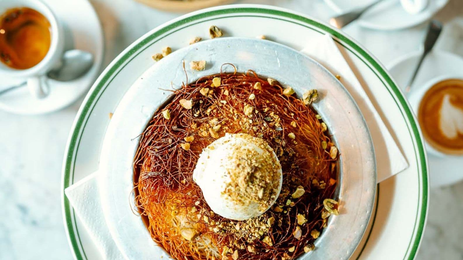 A plate of kunafa topped with ice cream and pistachios, surrounded by cups of coffee and another dessert on a marble table.