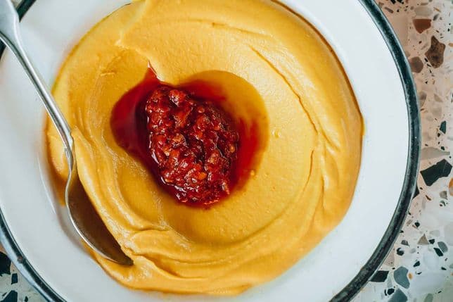 A plate of creamy hummus topped with a dollop of red sauce, served with a spoon on a terrazzo surface.