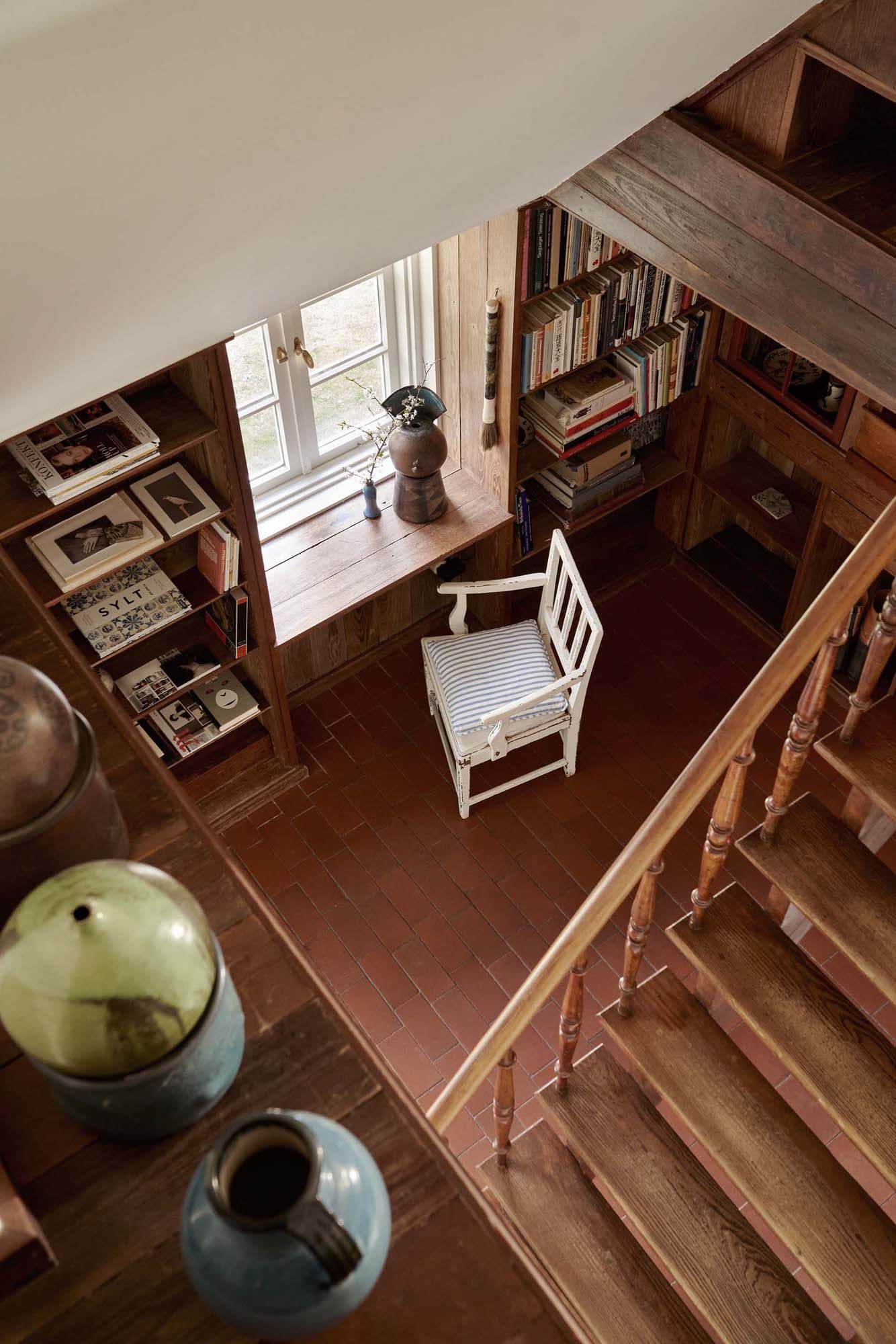 Cozy reading nook with a white chair, wooden shelves filled with books, a window, and decorative pottery on a brick floor.
