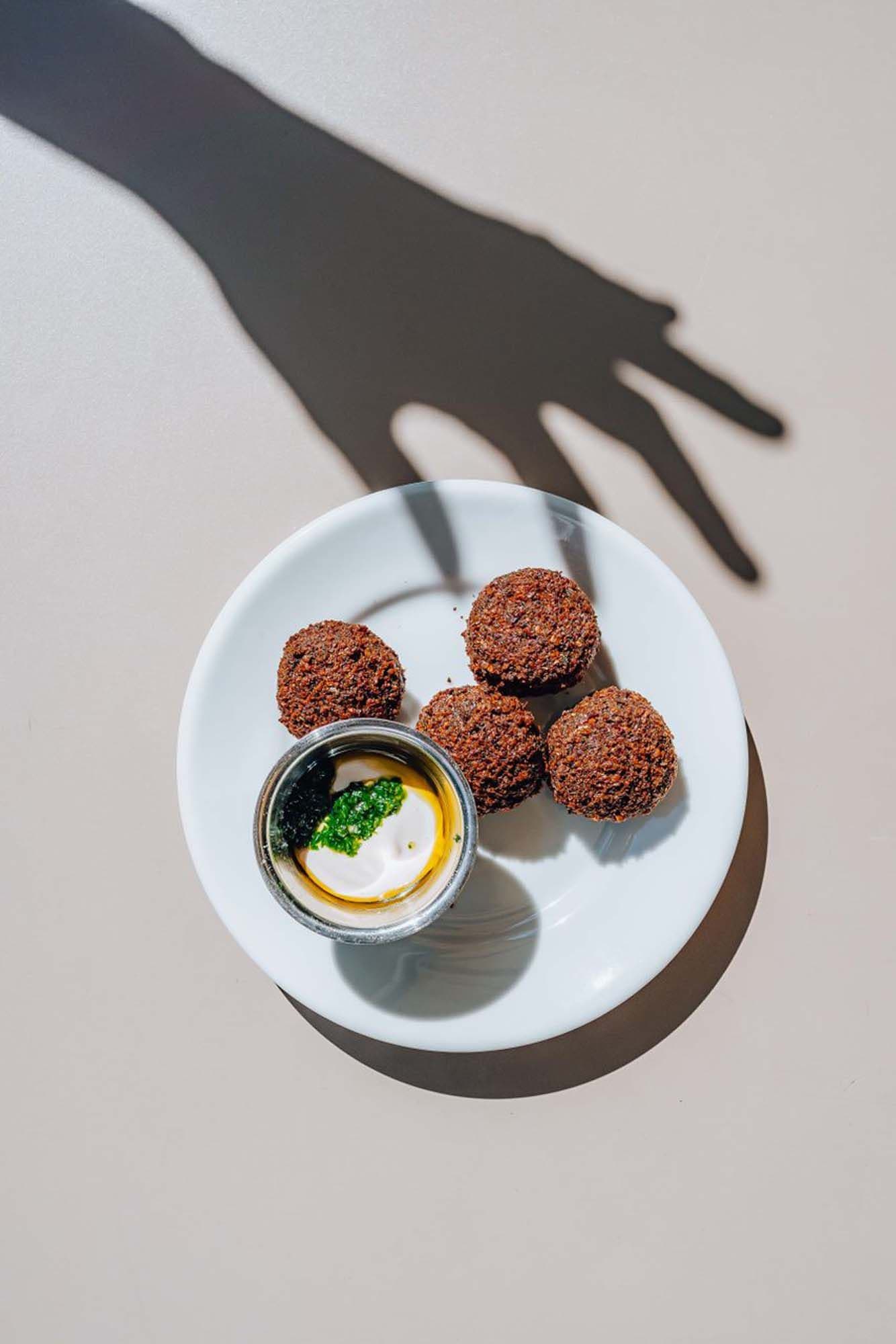 Plate with four falafel balls and a small bowl of dipping sauce on a white surface. A shadow of a hand is cast over the plate.