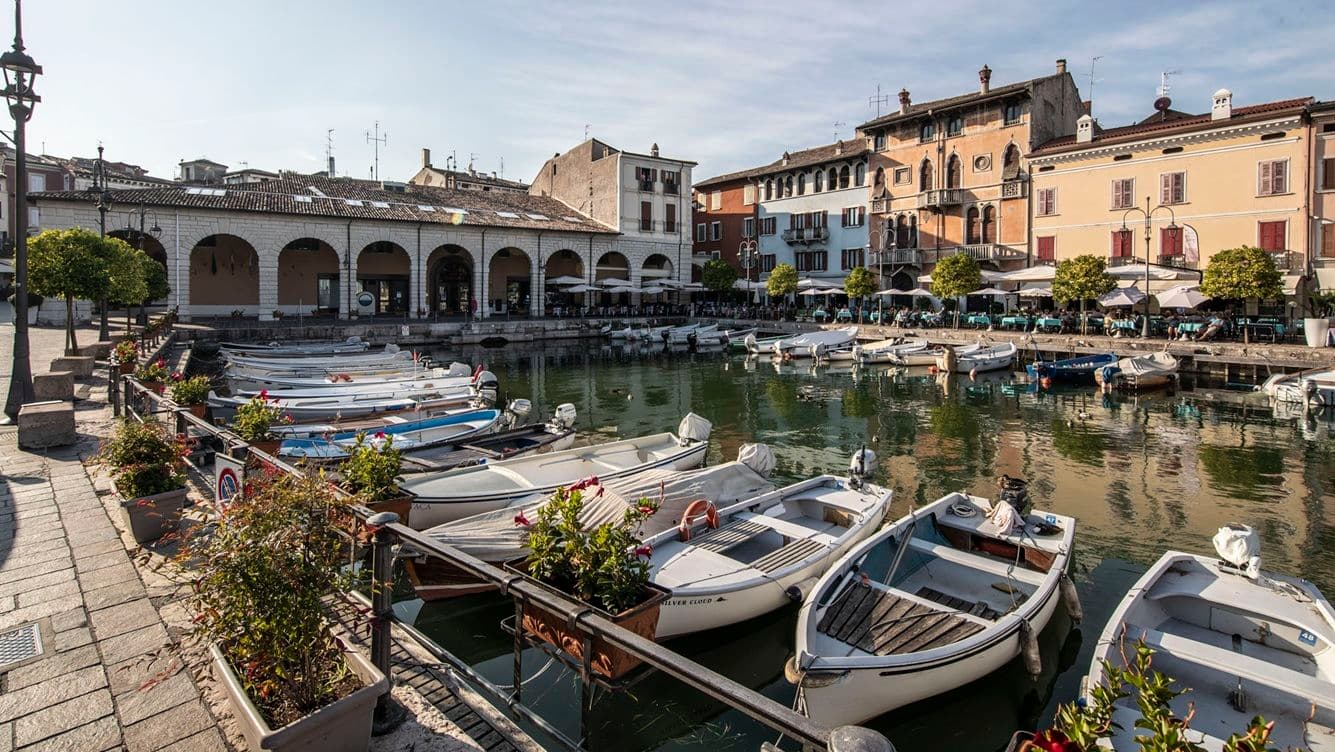 A scenic harbor with small boats docked, surrounded by historic buildings and a waterfront promenade lined with trees and flowers.