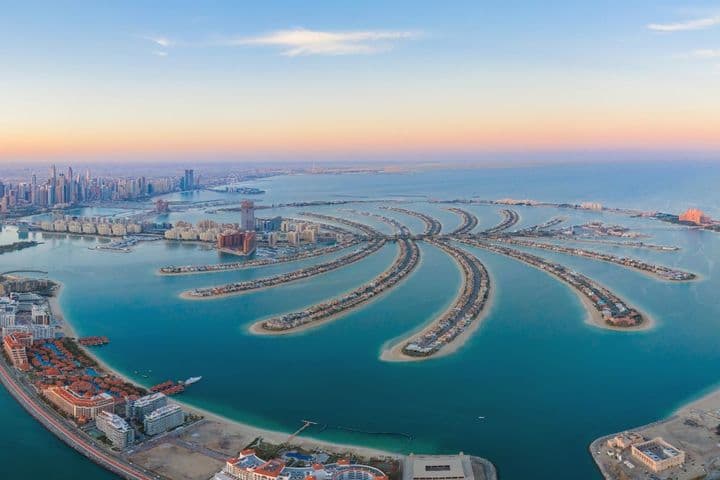 Aerial view of Dubai's Palm Jumeirah, a palm-shaped artificial archipelago, with city skyline and blue sea under a pastel sky.
