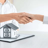 Duas pessoas apertam as mãos sobre uma secretária com uma pequena maqueta de uma casa, um contrato e uma caneta, simbolizando um acordo imobiliário.