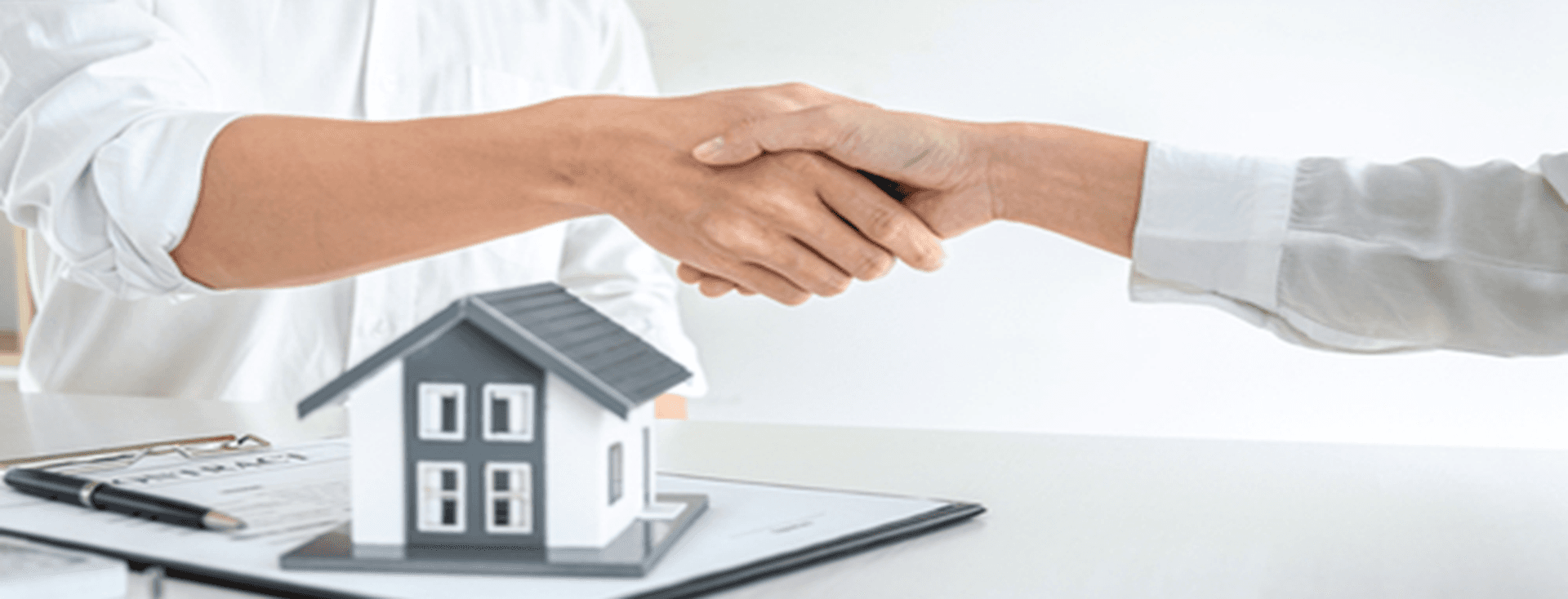 Zwei Personen, die sich über einem Schreibtisch die Hände schütteln, auf dem ein kleines Hausmodell, ein Vertrag und ein Stift liegen, symbolisieren eine Immobilienvereinbarung.