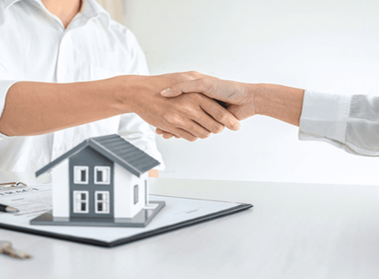 Zwei Personen, die sich über einem Schreibtisch die Hände schütteln, auf dem ein kleines Hausmodell, ein Vertrag und ein Stift liegen, symbolisieren eine Immobilienvereinbarung.