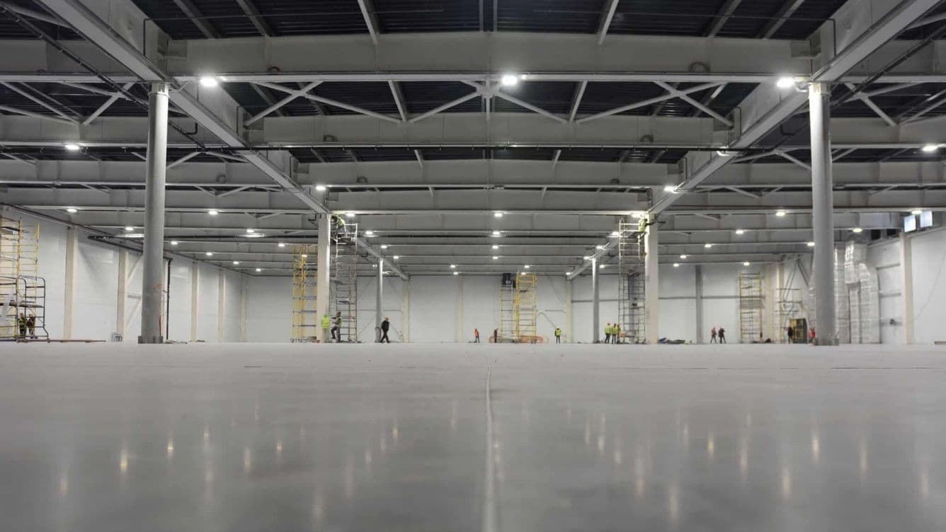 Interior espaiós d'una nau industrial amb sostres alts, bastides i terra de formigó. Es pot veure els treballadors de lluny.