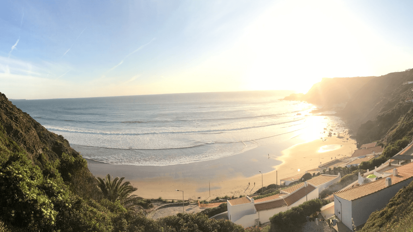 A panoramic view of a sunlit beach with gentle waves, surrounded by cliffs and greenery, under a clear blue sky.