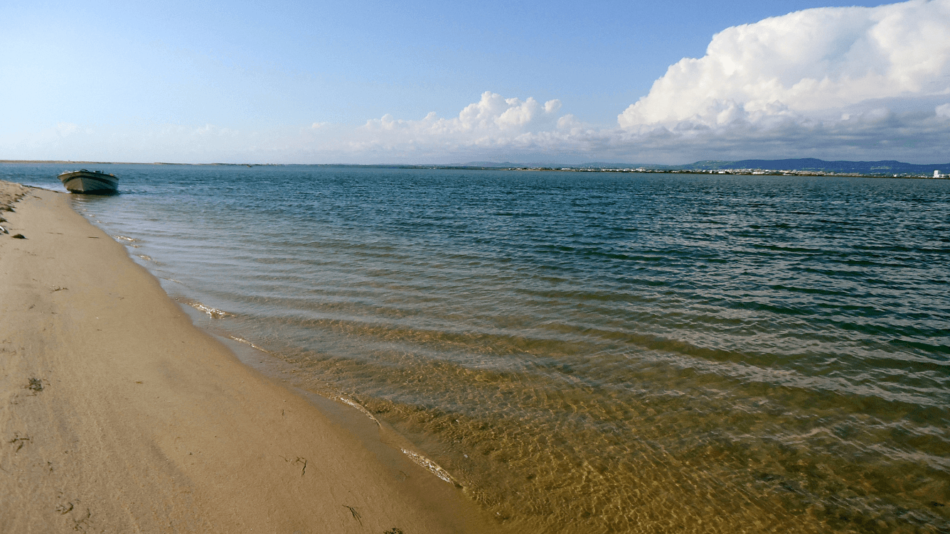 Sandy beach with a small boat on the shore, calm blue-green water, and a distant view of clouds and land on the horizon.