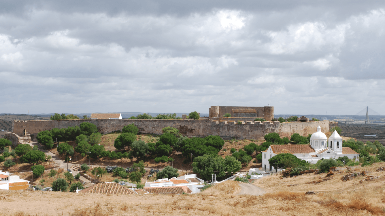 A historic fortress atop a hill with surrounding greenery, white buildings, and a distant bridge under a cloudy sky.