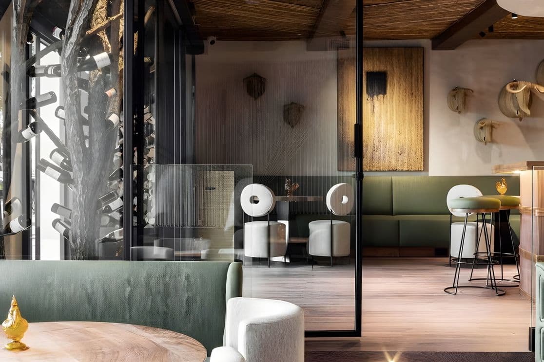 Modern restaurant interior with green seating, round white tables, wooden ceiling, abstract art, and decorative wall mounts. Glass partition in view.