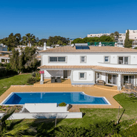 Vista aérea de uma moradia moderna branca com telhado em telha, ampla piscina e jardim exuberante, situada numa zona residencial sob um céu azul límpido.